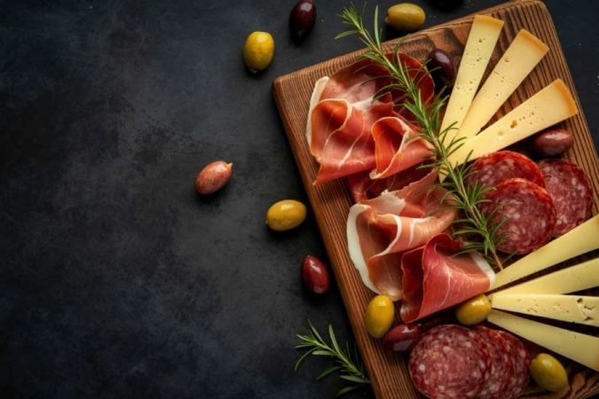 Planche apéritive pour un dîner gastronomique avec jambon cru, saucisson, fromage affiné, olives et romarin frais sur fond sombre.