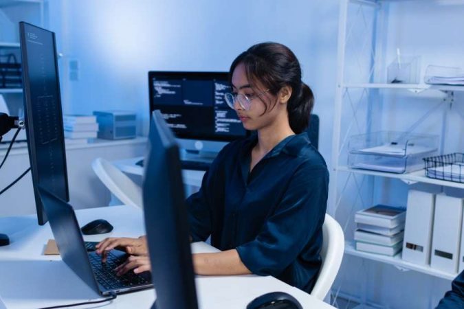 Une jeune femme à lunettes travaille sur son ordinateur portable au milieu d'un bureau équipé de plusieurs écrans pour maîtriser le prompting sur Gemini.
