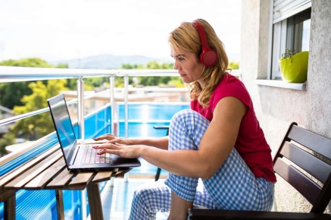 Une femme en pyjama et casque audio tape sur son ordinateur portable sur un balcon, illustrant les défis de la vie de famille et télétravail.
