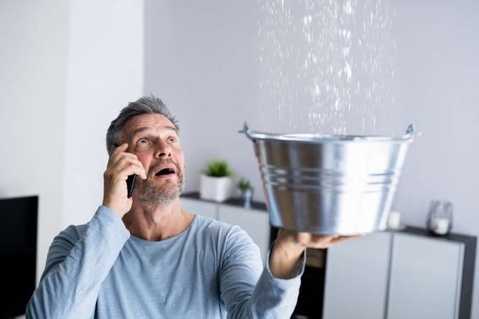 Un homme inquiet téléphone à un plombier tandis que l'eau de la cuisine s'écoule du plafond dans un seau.